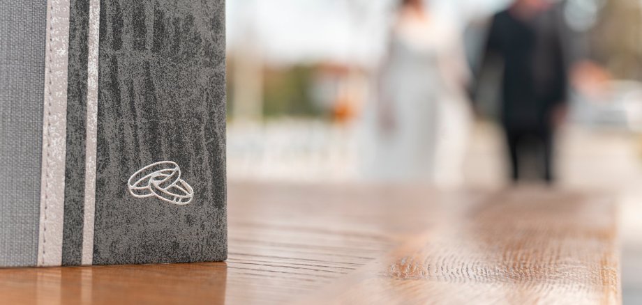 Weddings Grey family register with two wedding rings. A married couple in the blurred background.