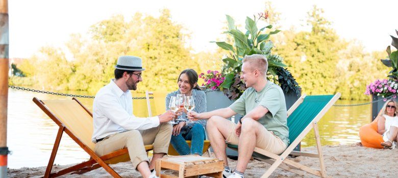 Two men are sitting with a woman on the beach at the Promenade Wine Festival, clinking their wine glasses.