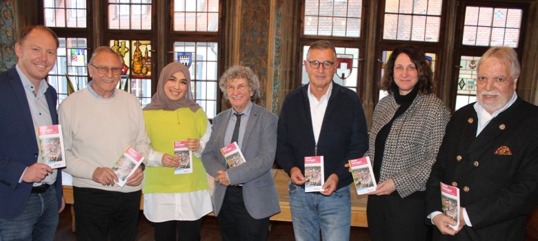 Lord Mayor Stefan Güntner, Kurt Semmler, Sümeyra Özkan, Dr Georg Feser, Chairman Ralph Hartner, Kerstin Baderschneider and Herrmann Reifenschied stand next to each other in the historic wedding hall, holding a flyer in their hands.