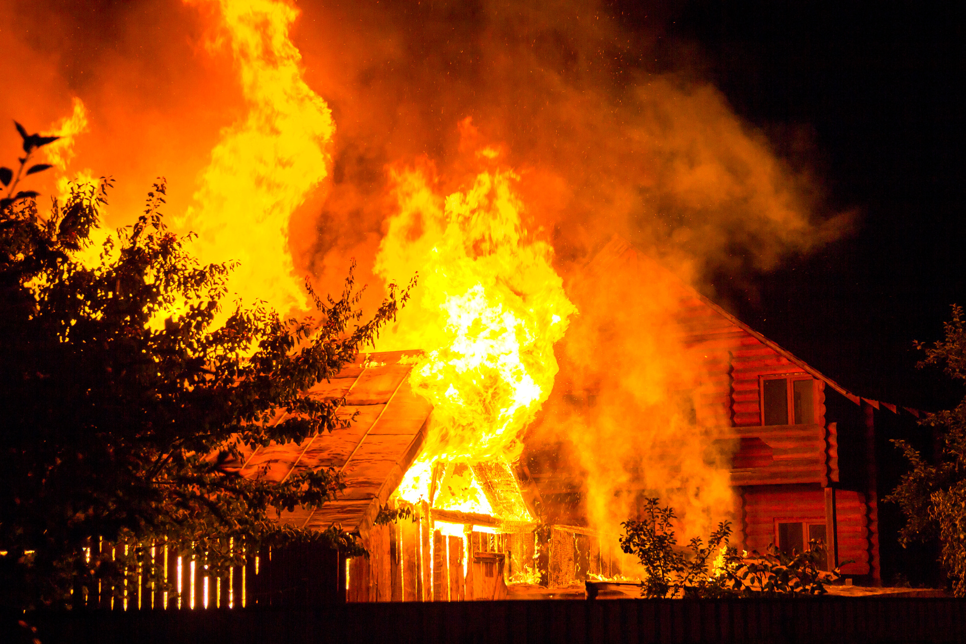 Brennendes Holzhaus bei Nacht Brennendes Holzhaus bei Nacht. Helle orangefarbene Flammen und dichter Rauch steigen unter dem Ziegeldach auf, im Hintergrund zeichnen sich Baumsilhouetten und die Häuser der Nachbarn ab.