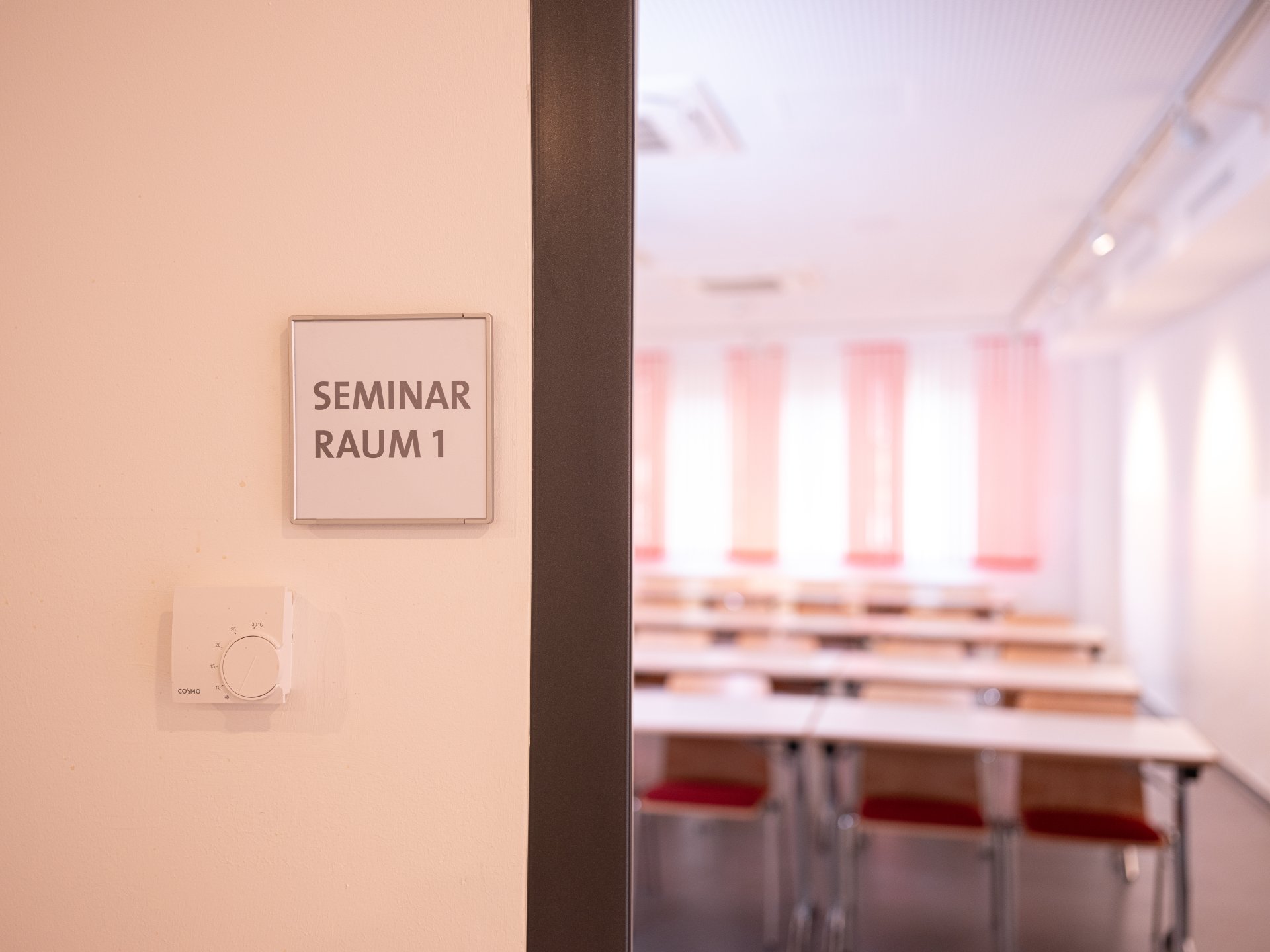 Seminar room Fastnachtakademie Open door with a view of tables and chairs standing in a row. There is a sign on the wall next to the door. It says "Seminar room 1"